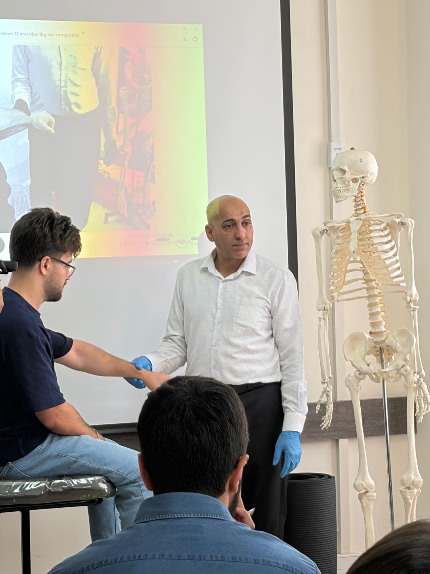 Dr. Hussein Nasser demonstrates a hand technique on a seated participant beside an anatomical skeleton model